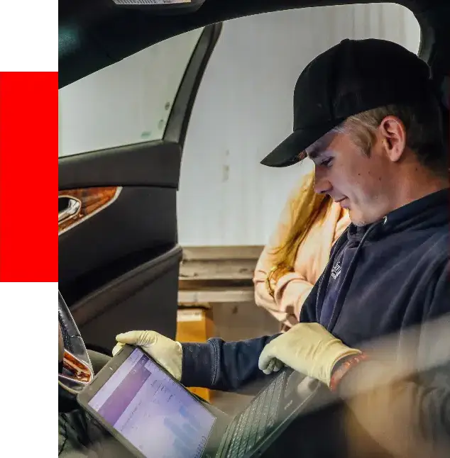 A person in a cap and gloves focuses on a laptop inside a car, possibly conducting preventative maintenance services, while another individual is partially visible in the background.