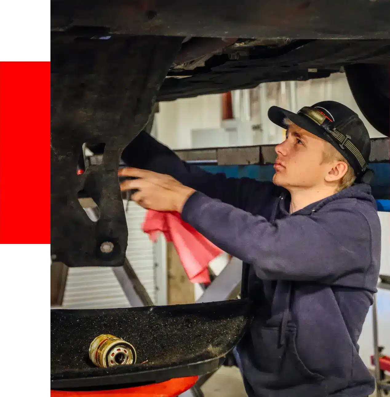A mechanic in a blue hoodie works underneath a car on a lift, holding a red cloth, expertly performing fluid maintenance services.